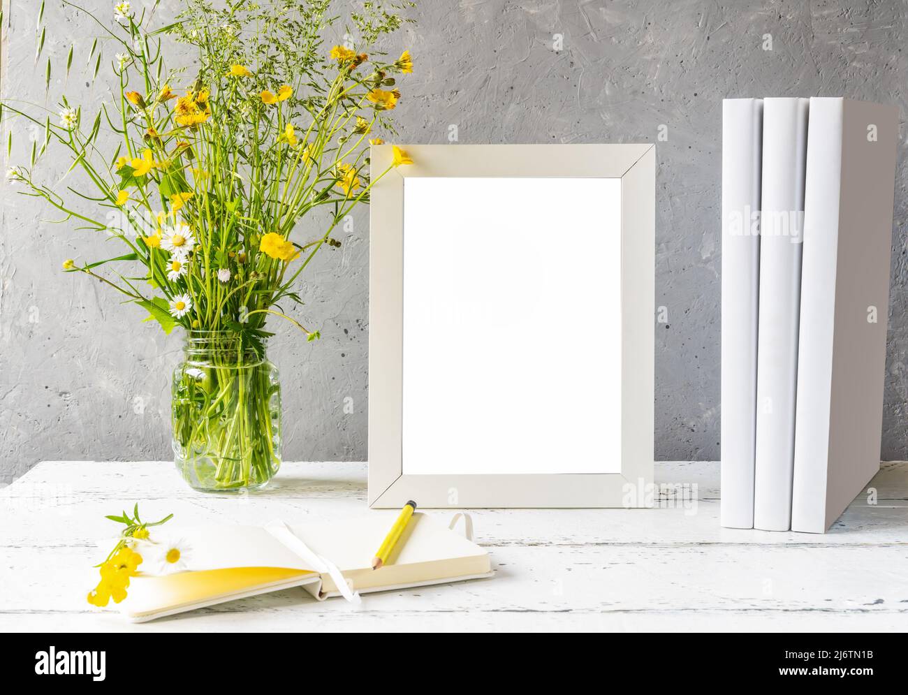 Modellerstellung. Weißer Rahmen auf weißem Holztisch mit grauem Betonhintergrund, Büchern, Notizbuch und wilden Blumen Stockfoto