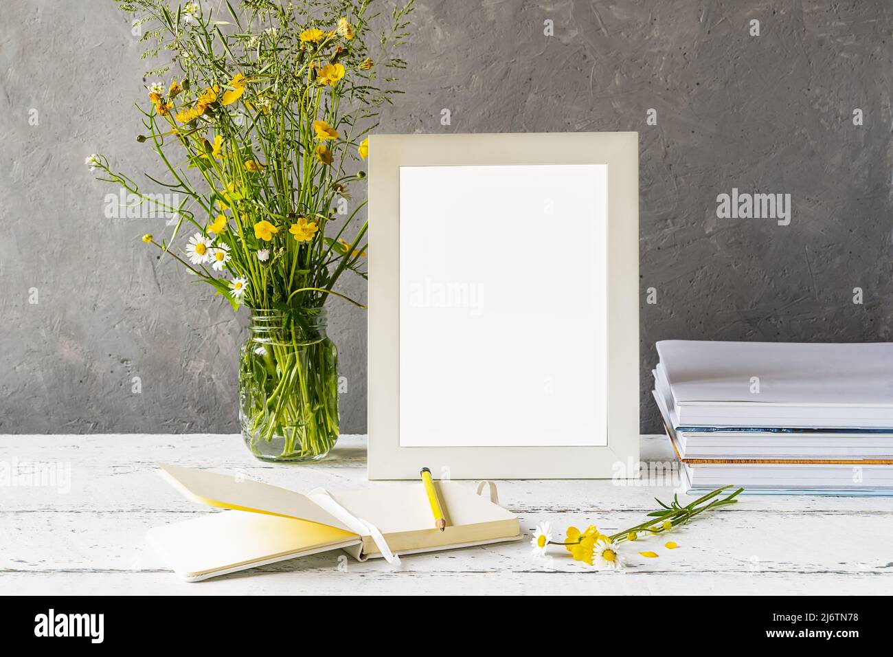 Modellerstellung. Weißer Rahmen auf Holztisch mit grauem Betonhintergrund, Büchern, Notizbuch, Bleistift und Wildblumen Stockfoto