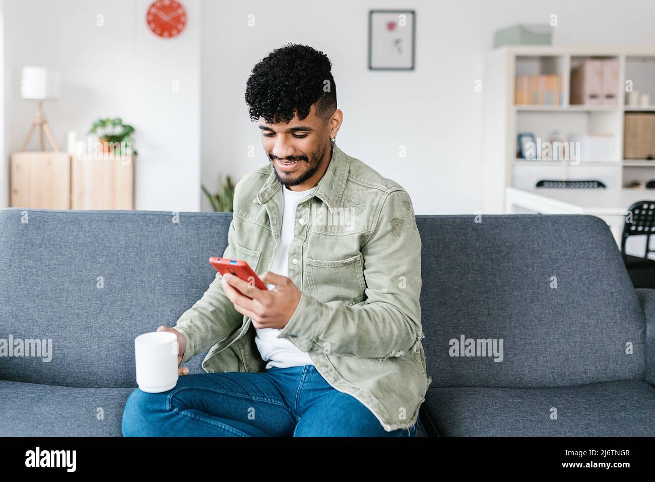 Junger Erwachsener, der zu Hause auf einem Sofa sitzt und ein Mobiltelefon benutzt Stockfoto