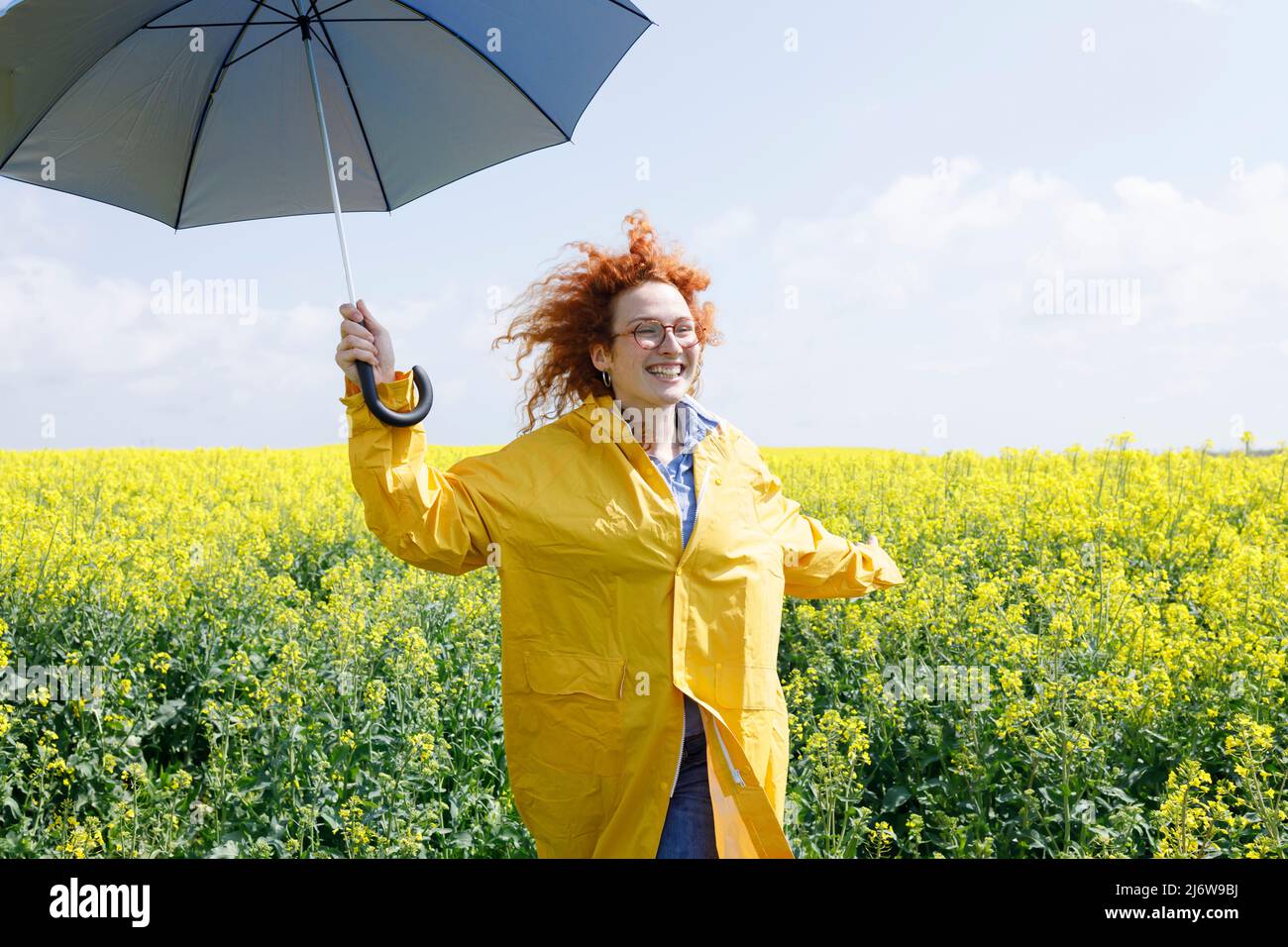Eine Frau, die mit dem Regenschirm durch ein Feld von gelbem Raps läuft und den sonnigen Sommertag genießt Stockfoto