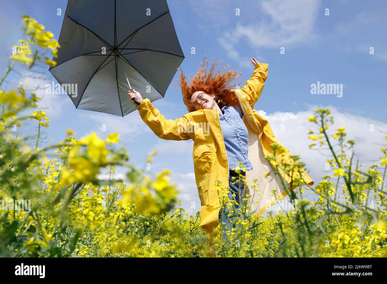 Eine Frau mit einem blauen Hemd und einem gelben Regenmantel, die auf dem Feld des gelben Raps hüpft und sich an einem sonnigen Sommertag amüsieren kann Stockfoto
