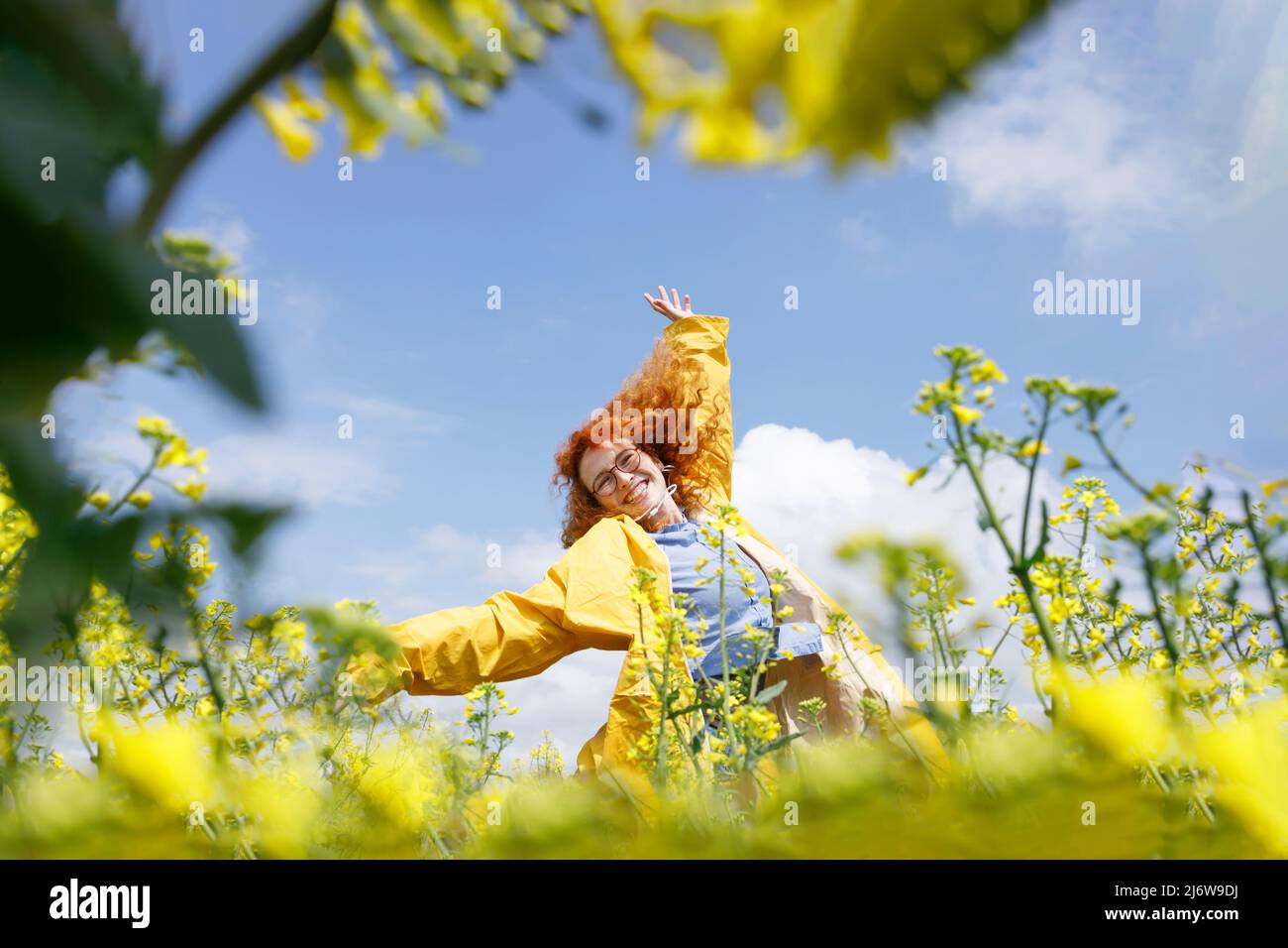 Eine Frau in einem gelben Regenmantel, die auf dem Feld der gelben Blumen springt und an einem sonnigen Tag Spaß hat Stockfoto