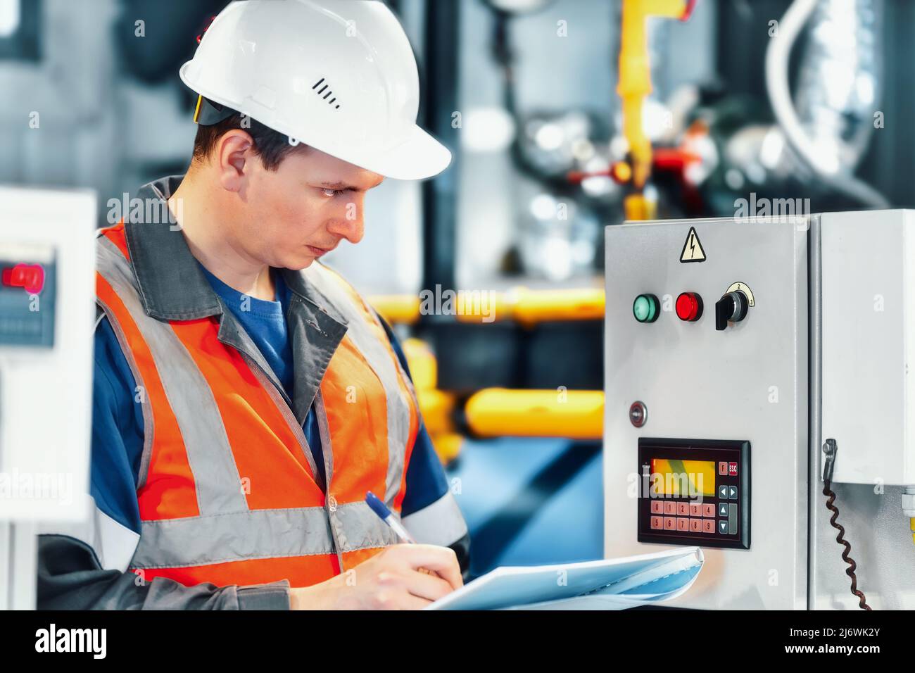 Der junge Ingenieur, der Helm und Schutzweste verwendet, zeichnet die Messwerte der Geräte oder des Gasverbrauchs im Kesselraum auf. Beginn der Heizperiode und Gasversorgung. Stockfoto