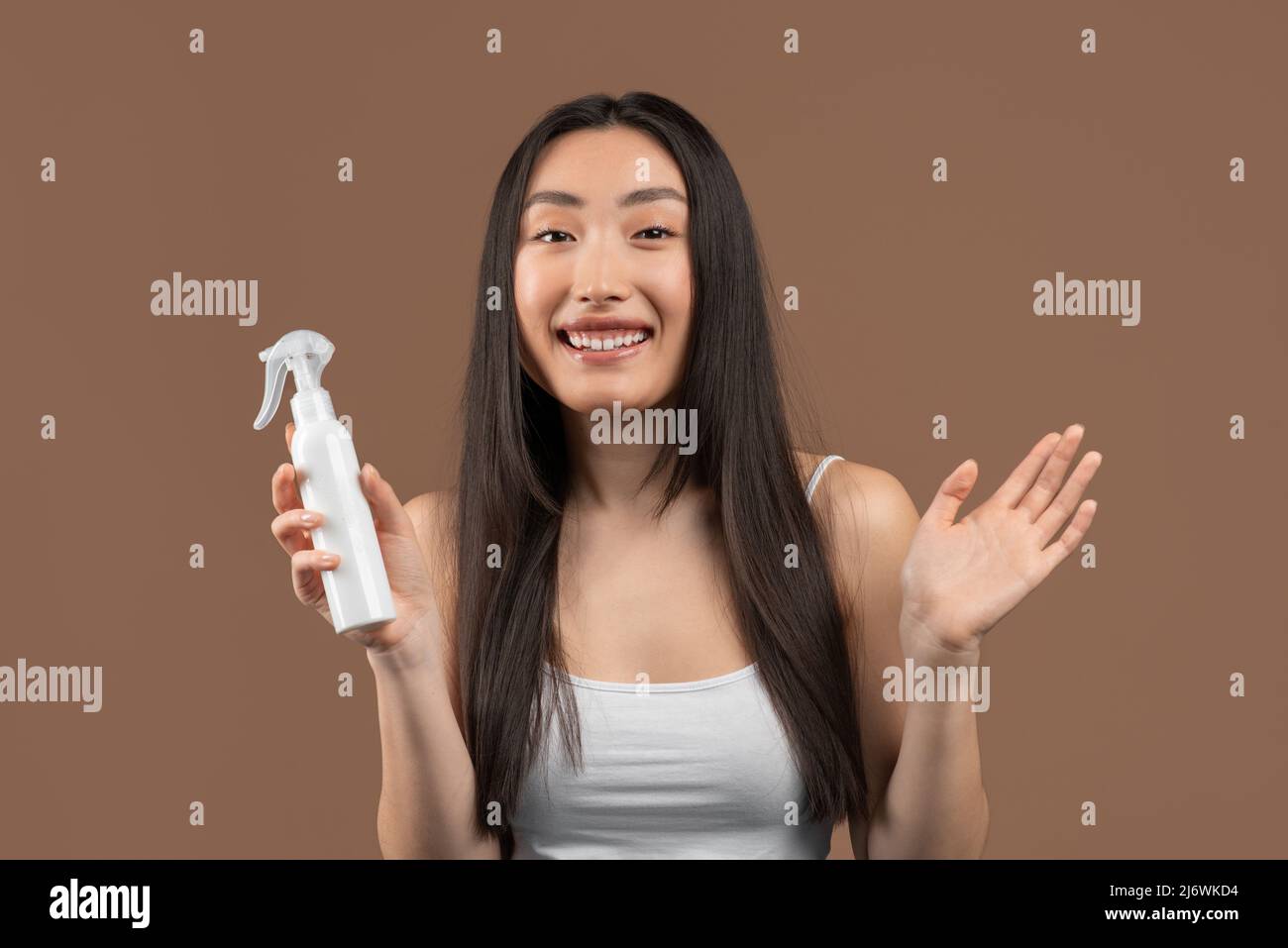 Hübsche koreanische Frau mit langen Haaren empfehlen Flasche mit Haarprodukt, Serum oder Öl, brauner Hintergrund Stockfoto