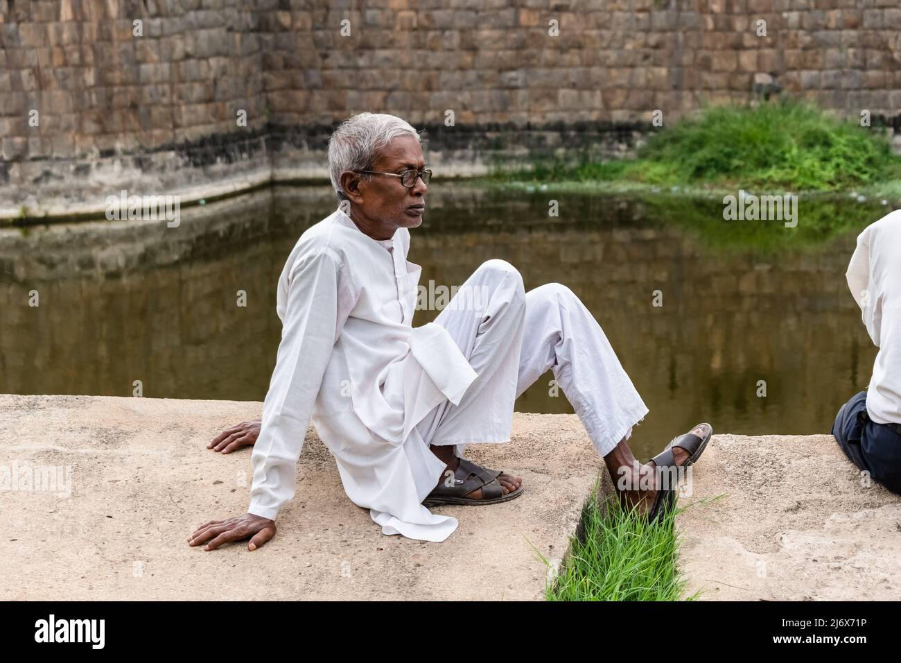 Vellore, Tamil Nadu, Indien - 2018. September: Ein älterer indischer Mann, der ein weißes Hemd und einen Pyjama trägt, sitzt allein am Rand einer Wand in Vellore Stockfoto