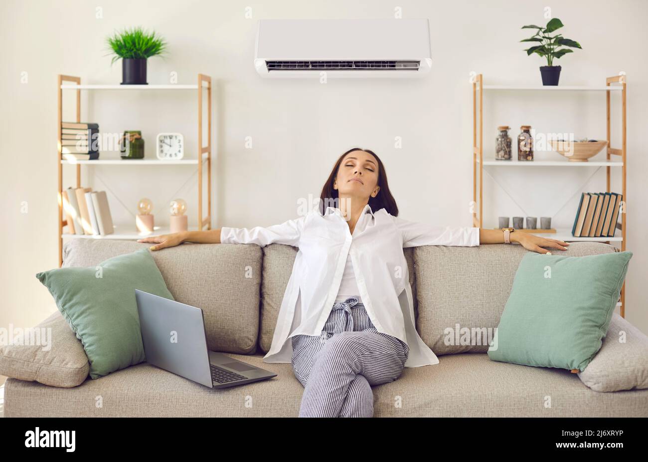 Entspannte Frau atmet frische Luft ein, während sie auf dem Sofa im Wohnzimmer mit Klimaanlage sitzt Stockfoto