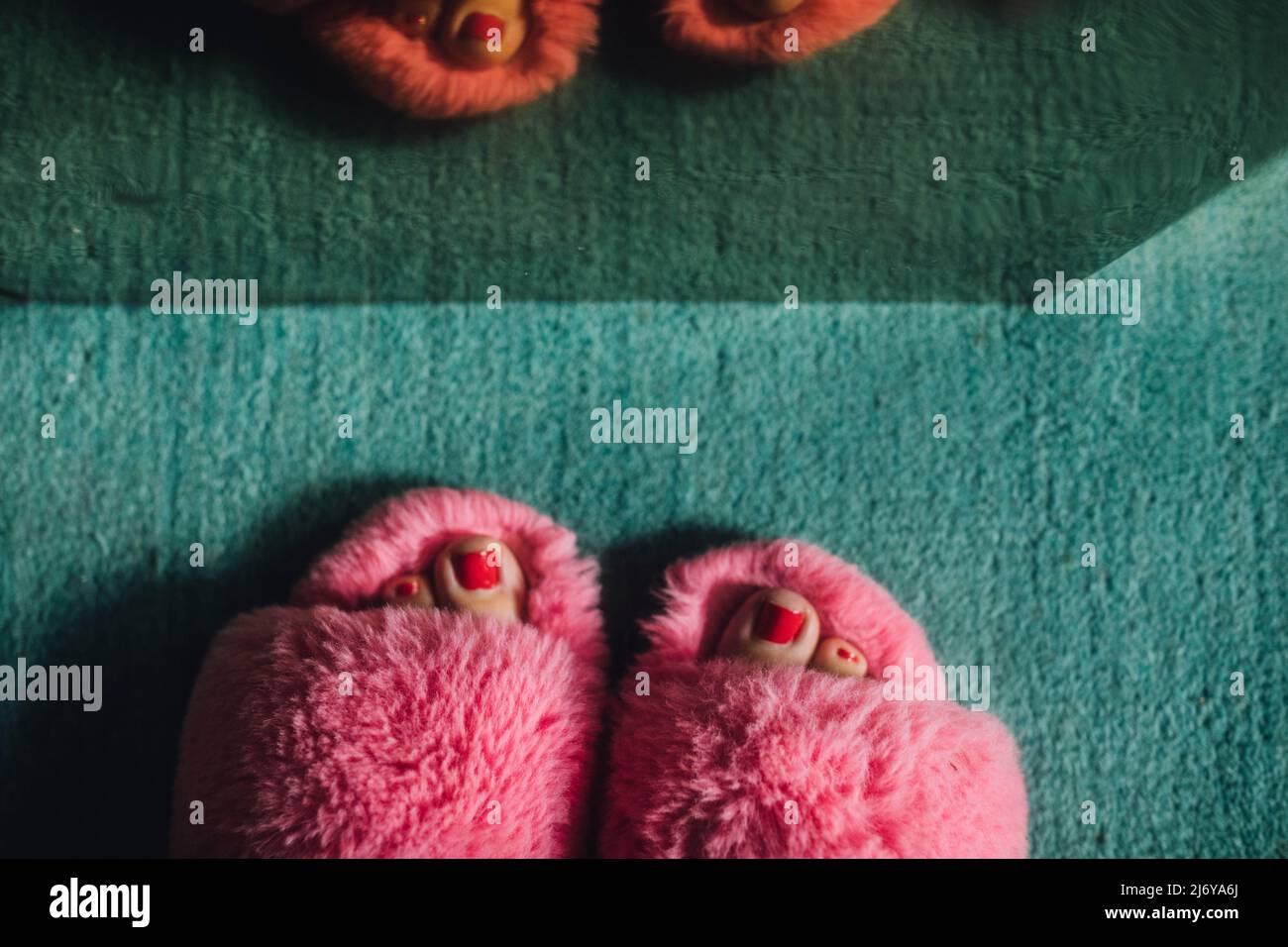 Füße mit rot gestrichenen Zehennägeln in rosa pelzigen flauschigen Pantoffeln gegen den tealfarbenen Teppich Stockfoto