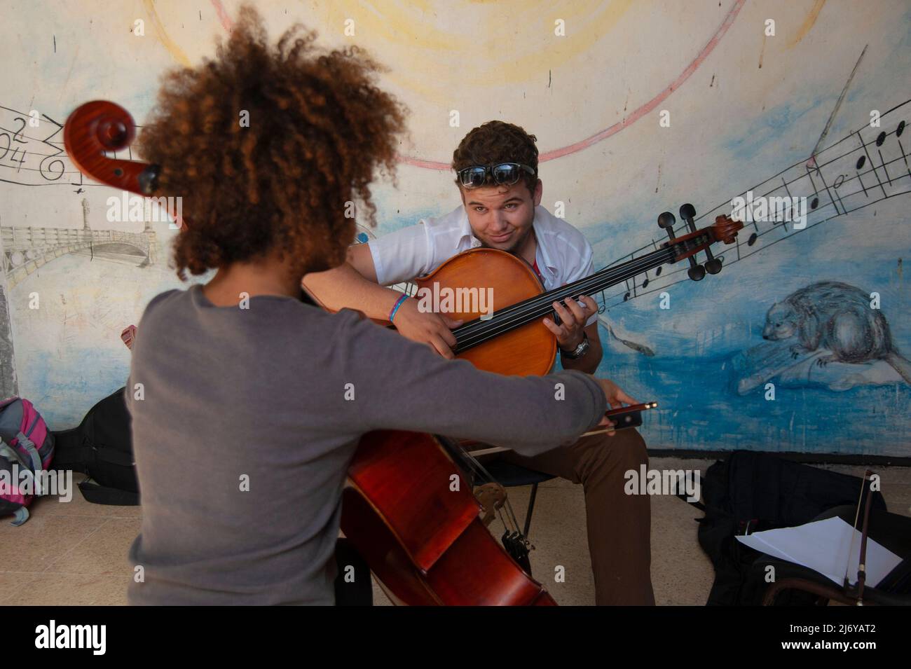 Junge Schülerinnen und Schüler Proben Cello an einer Schule in Matanzes, Kuba, mit Musikinstrument an der Wand und Wandbild dahinter. Stockfoto