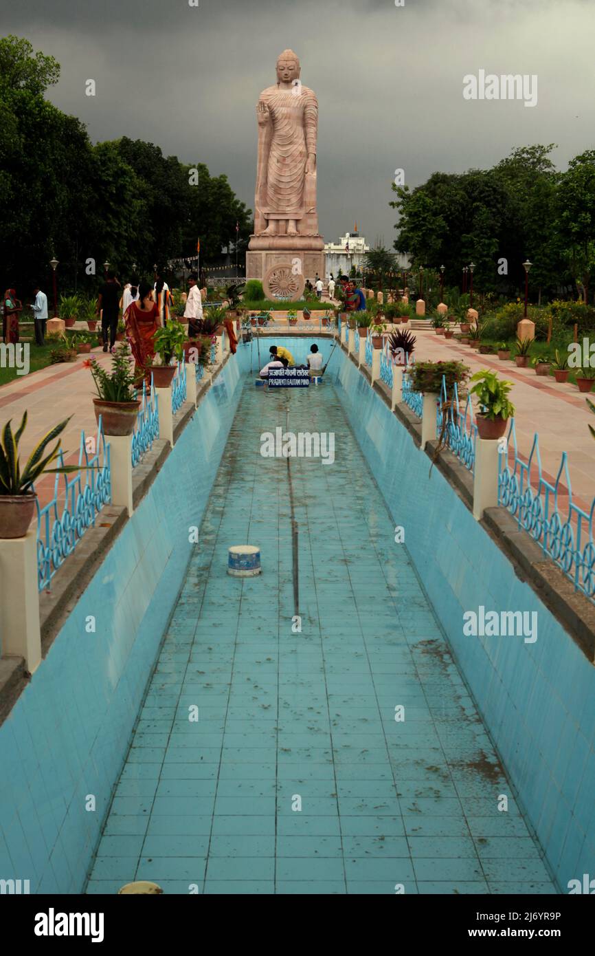 Ein Teich befindet sich im Vordergrund einer 80 Meter hohen Sandsteinstatue des stehenden Buddha in Sarnath am Stadtrand von Varanasi, Uttar Pradesh, Indien. Die von 1997 bis 2011 erbaute Statue war das Ergebnis einer gemeinsamen Anstrengung Thailands und Indiens. Stockfoto