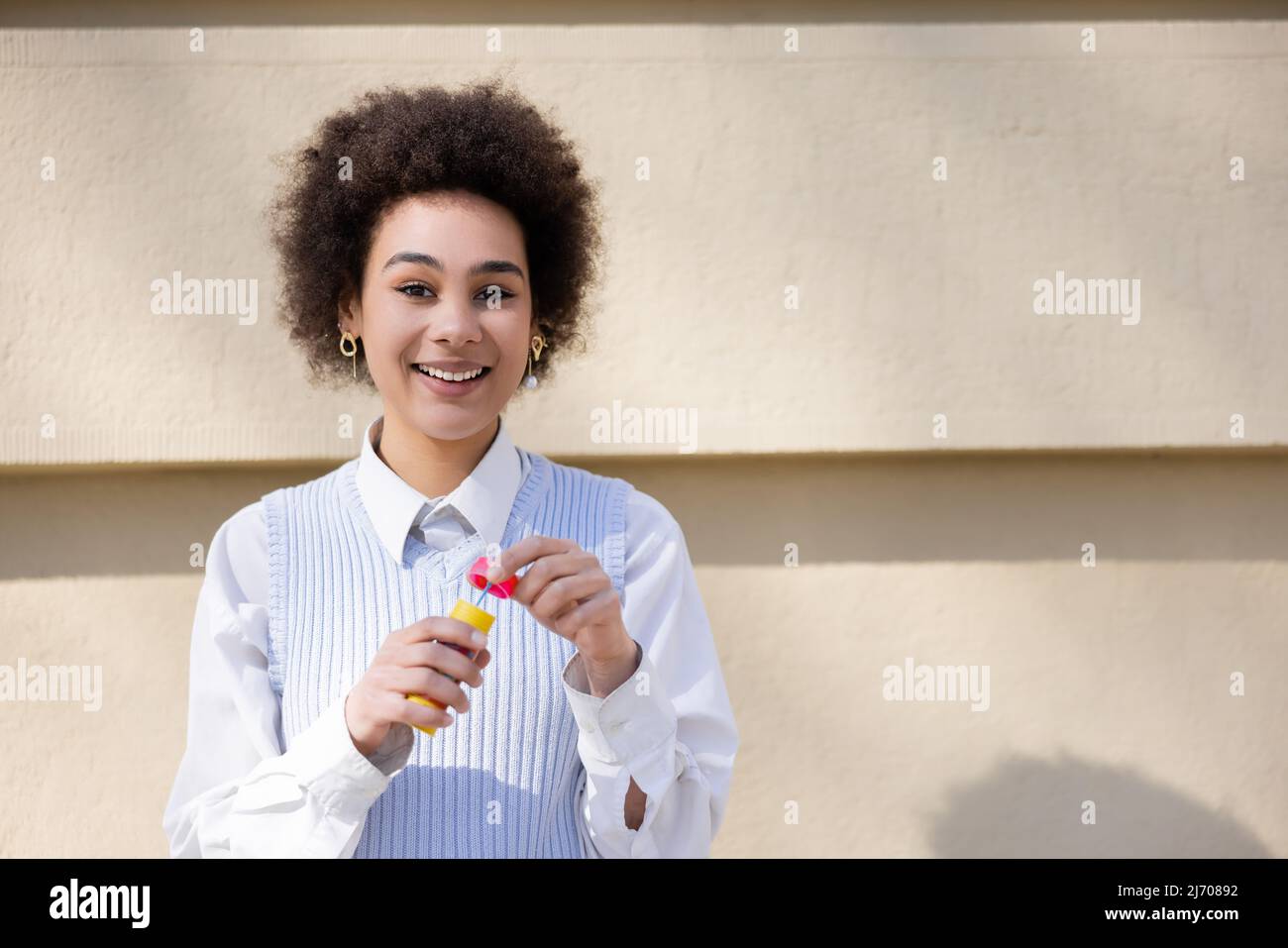 Glückliche afroamerikanische Frau in blauer Weste und weißem Hemd mit Flasche mit Seifenblasen Stockfoto