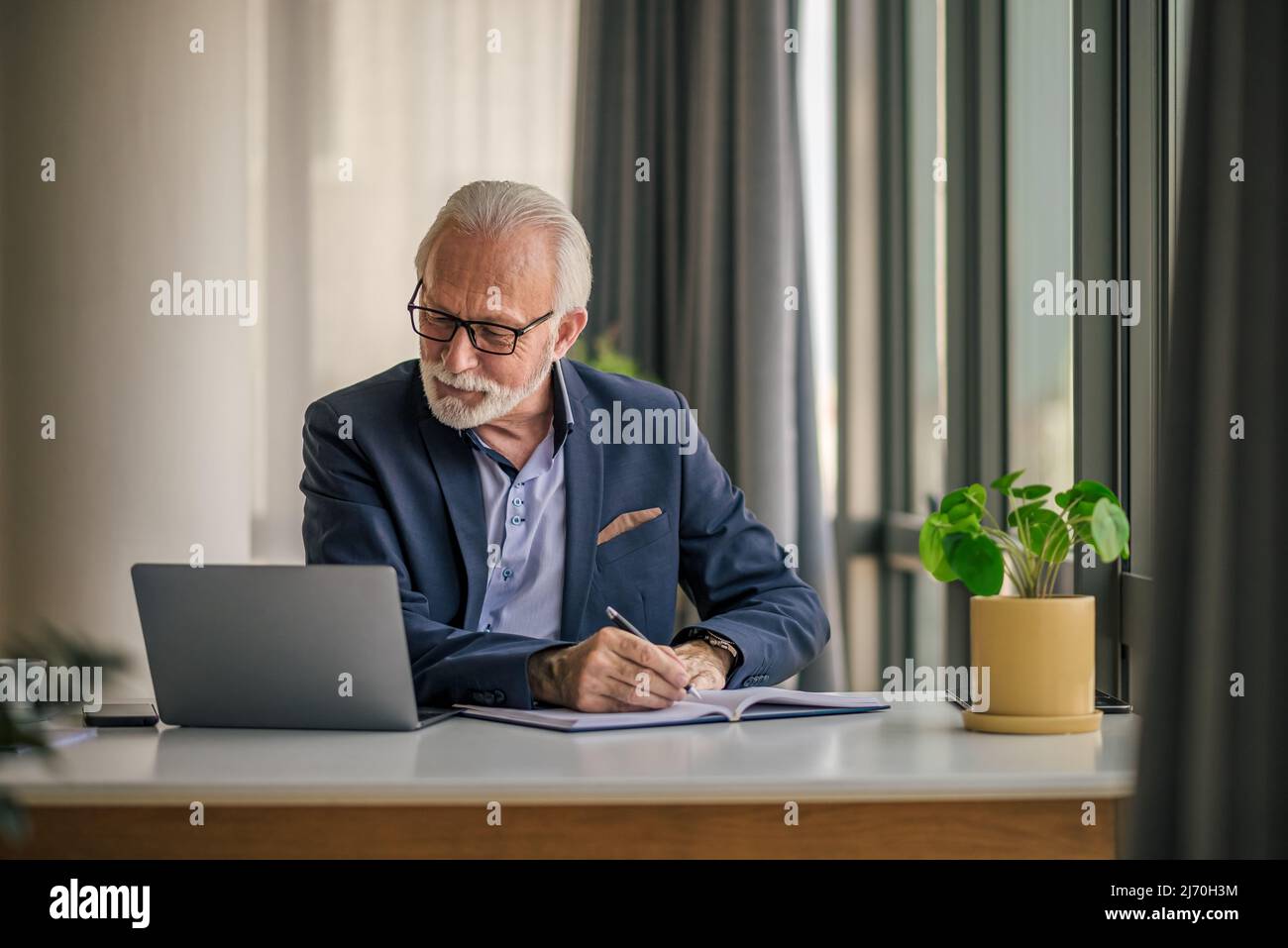 Leitender männlicher Profi, der an der Rezeption Tagebuch schreibt, älterer Manager, der einen Geschäftsplan erstellt, er benutzt einen Laptop, während er im Büro des Unternehmens arbeitet. Stockfoto