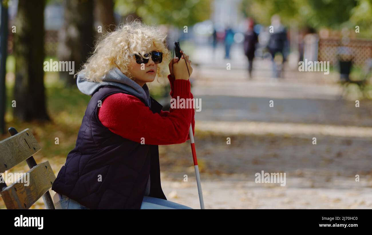 Die junge kaukasische blonde Frau mit Sehbehinderung sitzt auf einer Bank in einem Park und hält ihre Hände auf weißem Stock. Leben mit Behinderung Konzept. Hochwertige Fotos Stockfoto