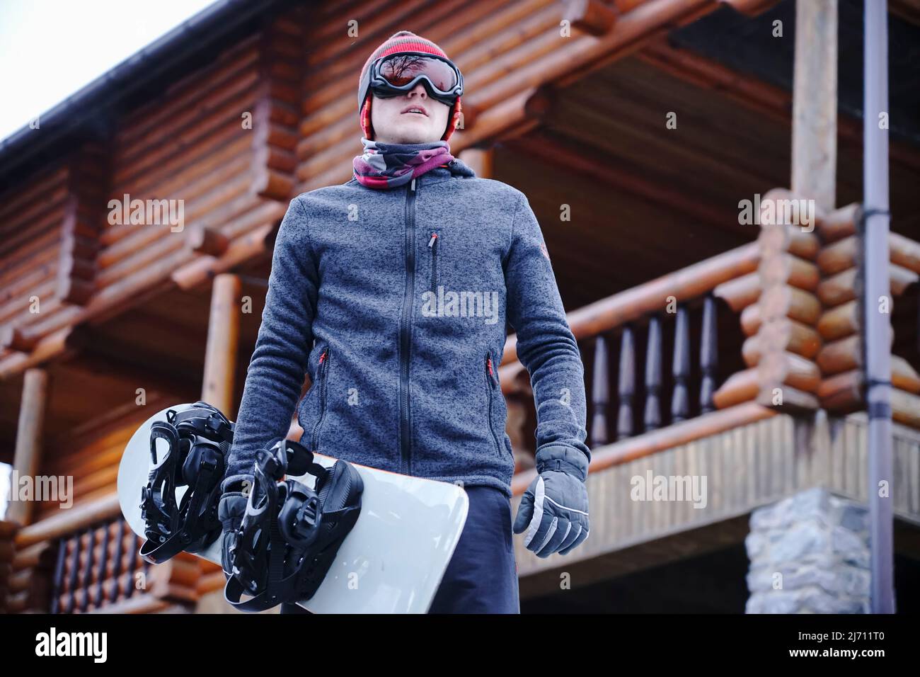 Ein Typ in Skibrille hält ein Snowboard in der Hand in der Nähe des Resorthauses in der Skistation. Hochformat Stockfoto