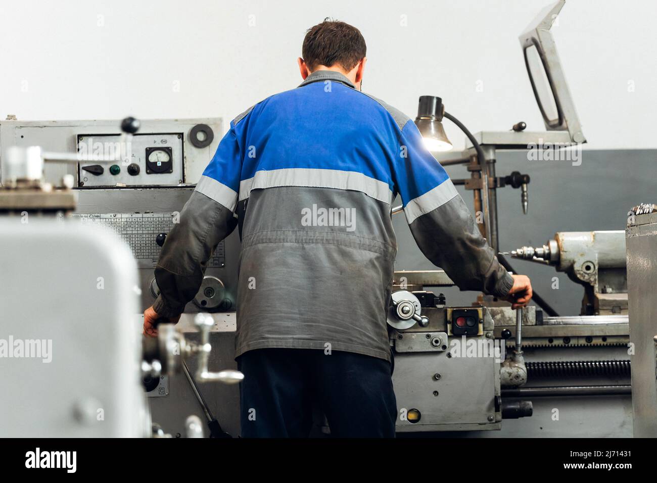 Turner steht hinter der Drehmaschine in der Produktionshalle und arbeitet. Blick auf den Arbeiter von hinten in Overalls. Authentische Arbeitsprozess-Szene in der Produktion. Echter Arbeiter. Nicht inszenierte Szene. Stockfoto