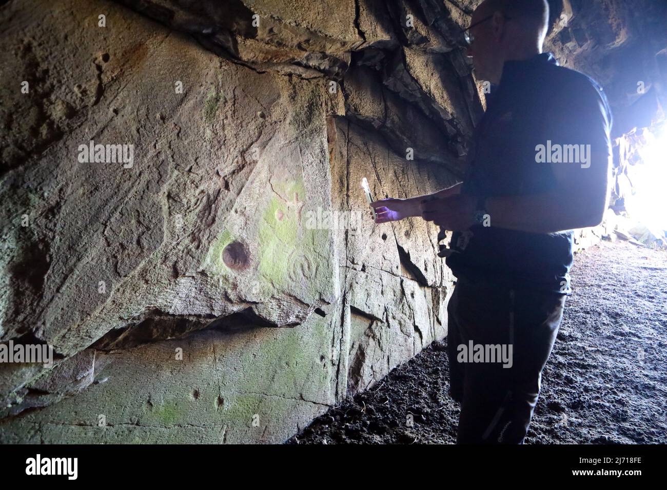 Ein Mann, der eine Fackel leuchtet, die die frühchristlichen Felszeichnungen und mögliche bronzefarbene Tassenmarkierungen in der Scoor Cave auf der Isle of Mull, Schottland, beleuchtet Stockfoto