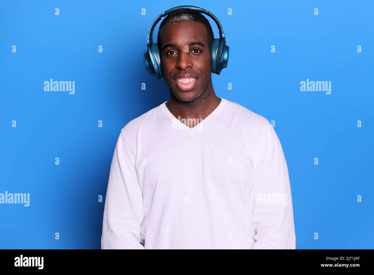 Porträt des Millennials Mann trägt Kopfhörer, lächelt breit, genießt Lieblingsmusik, gekleidet in weißen Pullover, isoliert auf blauem Hintergrund, hat glücklich Stockfoto