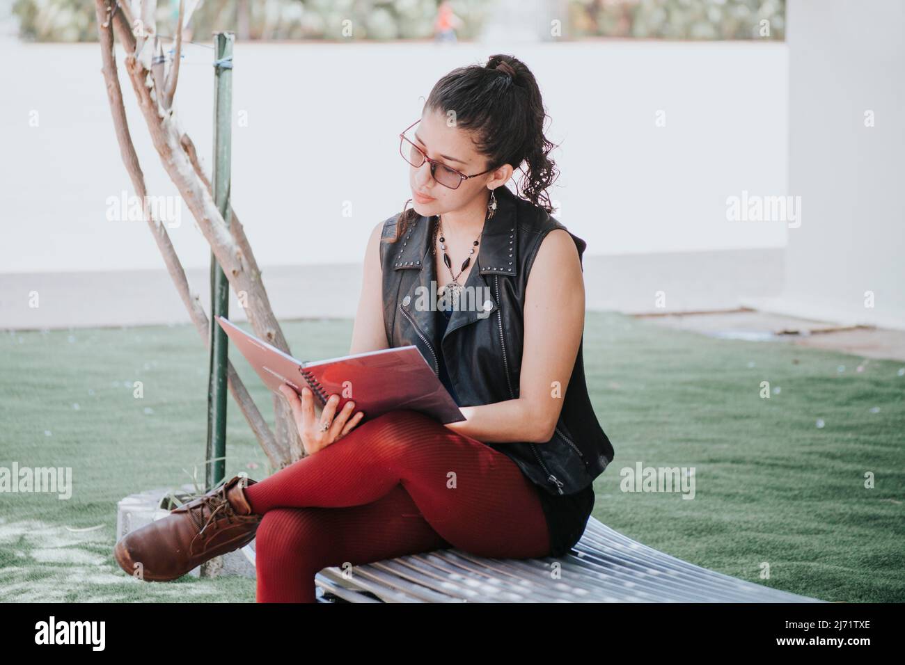 Ein Studentinnen liest ein Buch, ein Stadtmädchen sitzt auf einer Bank und liest ein Buch, ein Lateinerin liest ein Buch draußen Stockfoto