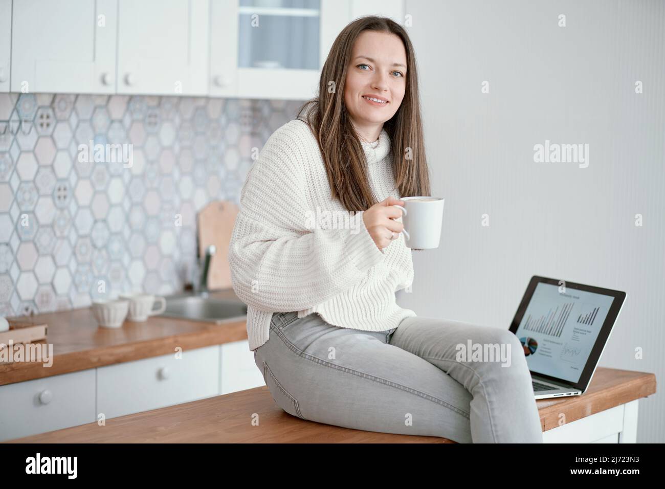 Junge Frau mit einer Tasse Kaffee, die auf ihren Laptop-Bildschirm schaut. Stockfoto