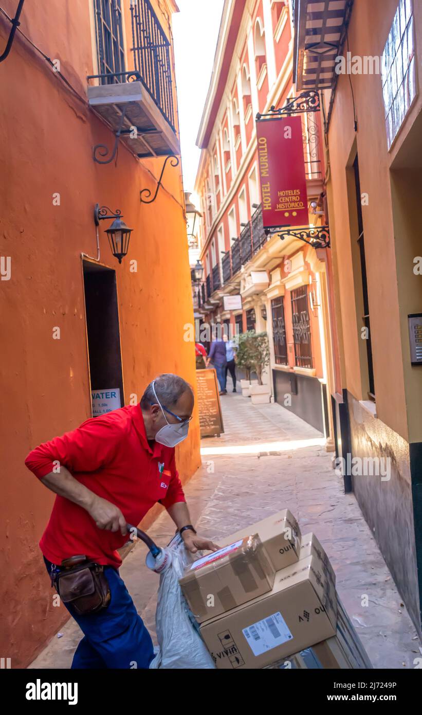 Delivery man in Gesichtsmaske verwaltet, um Boxen auf einer rollenden Karte in der engen Straße im Zentrum von Sevilla, Andalusien, Spanien zu liefern Stockfoto