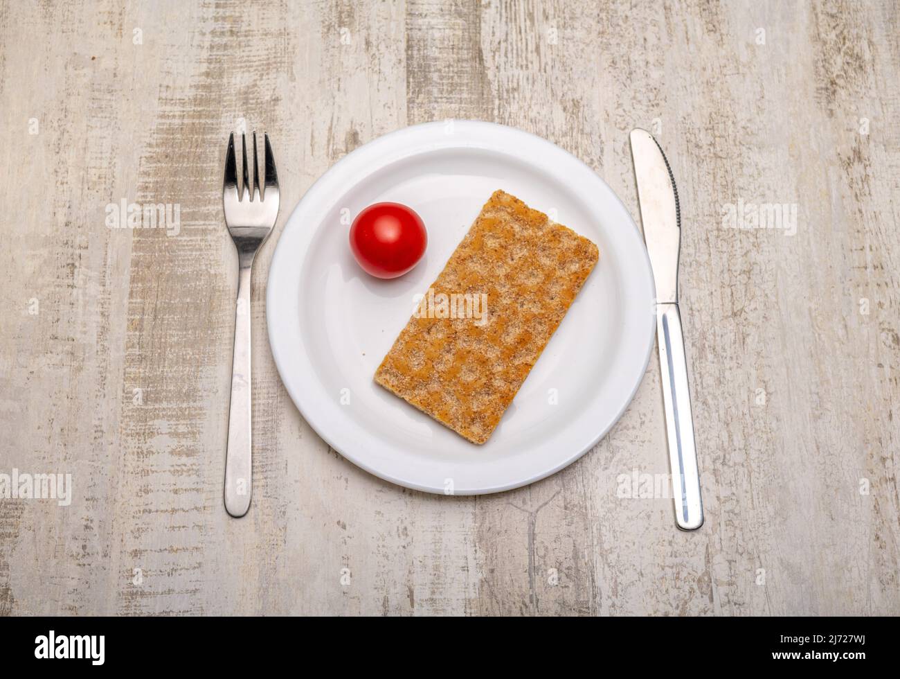 Eine einzelne Tomate und eine Scheibe Knäckebrot auf einem weißen Teller und Besteck mit Gebrauchsspuren auf einer rustikalen Holzoberfläche Stockfoto