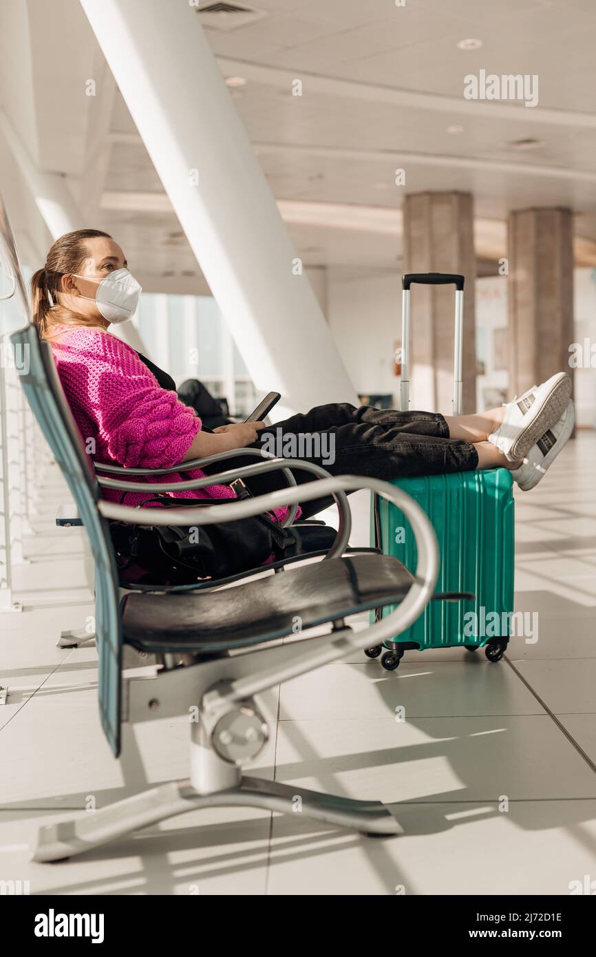 Vertikale Frau in Maske mit Gepäck sitzen im Wartezimmer mit Smartphone am internationalen Flughafenterminal. Coronavirus Stockfoto