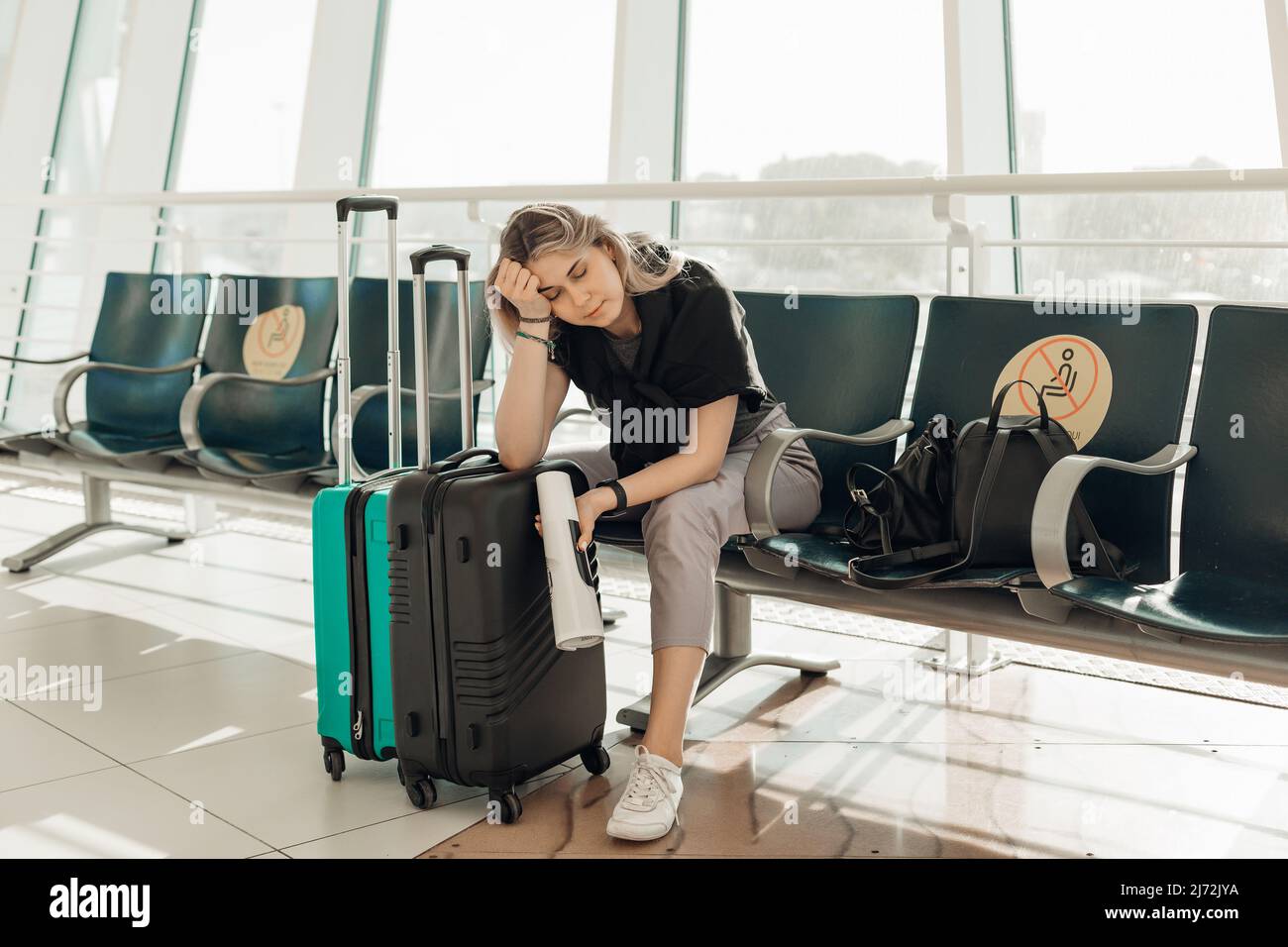 Gelangweilte blonde Frau mit Gepäck, Ellenbogen auf Taschen lehnend, sitzen im Wartezimmer am Flughafen wegen Reisebeschränkungen von Covid Stockfoto