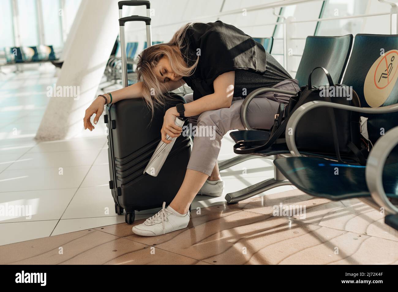 Müde und müde blonde Frau in legerer Kleidung mit Gepäck, auf Taschen gelehnt, sitzt im Flughafenterminal. Flug stornieren Stockfoto