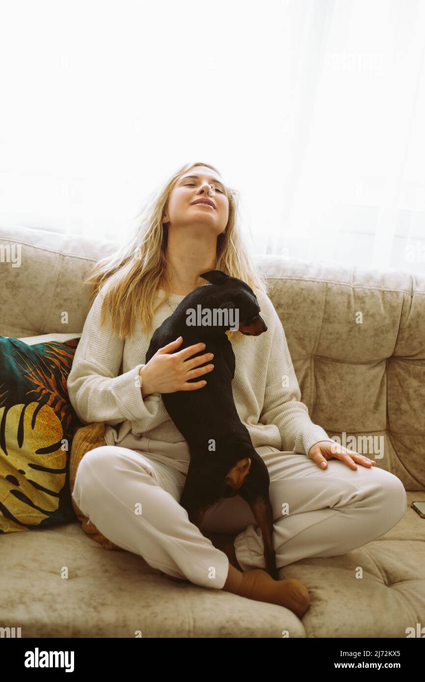 Vertikales Foto einer blonden Frau, die ihren besten Freund, den schwarzen Hund, auf dem Sofa umarmt. Entspannen und kuscheln. Kommunikation mit Tieren Stockfoto