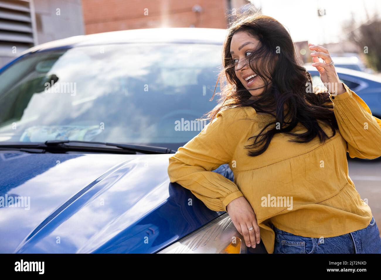 Lächelnde junge Frau, die über ihre Schulter blickt, mit ihren im Wind wehenden Haaren, die sich gegen das Auto lehnen. Stockfoto