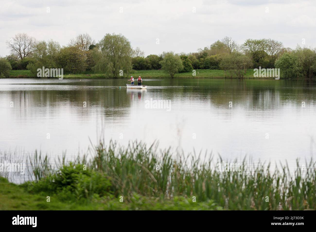 Zwei männliche Angler stehen auf und werfen eine Linie / gehen Angeln in einem kleinen Ruderboot in der Mitte eines Sees / Wasser, Großbritannien Stockfoto