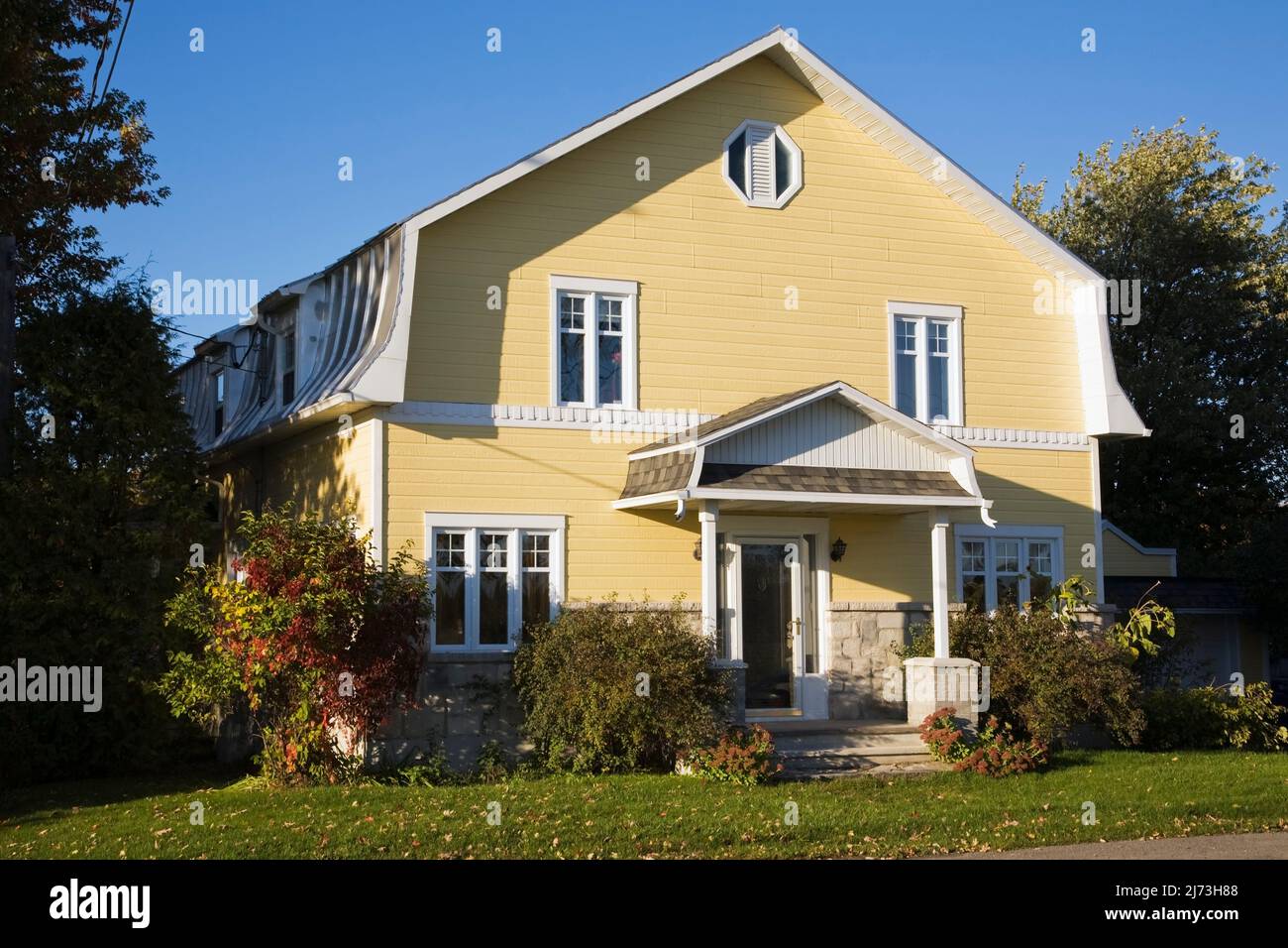 Gelbes zweistöckiges Haus mit Stein- und Holzplanken im Scheunenstil mit landschaftlich gestaltetem Vorgarten im Herbst. Stockfoto