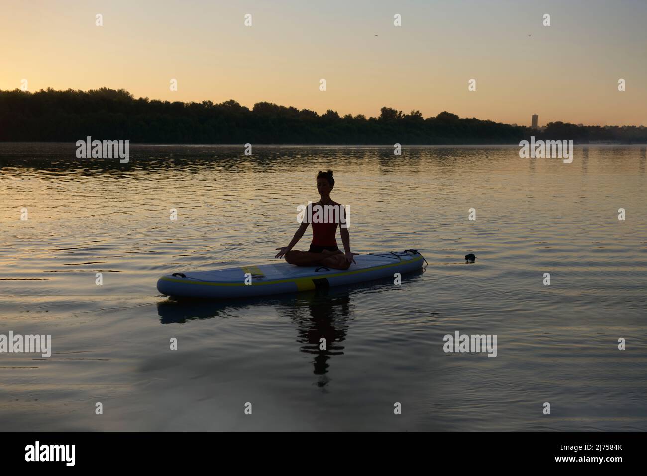 Silhouette einer Frau, die am frischen und warmen Sommermorgen auf einem Paddle-Board sitzt und meditiert Stockfoto