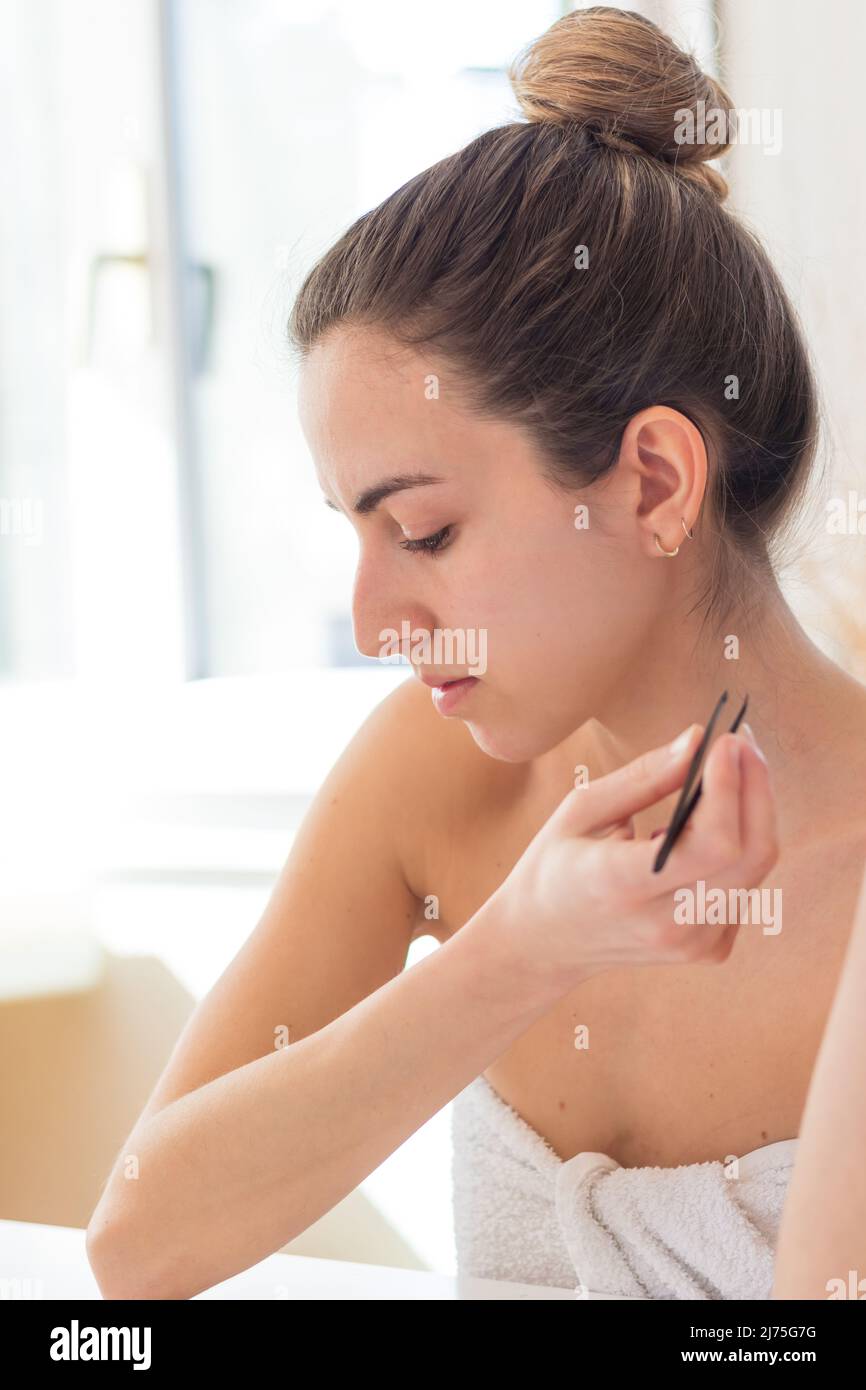 Junge Frau gerade aus der Dusche mit Brötchen und Handtuch auf ihrem Körper und einer Pinzette in der Hand, die nach unten schaute Stockfoto