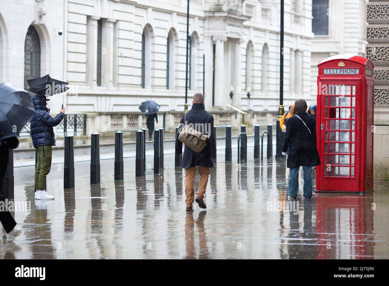 Eine Person, deren Regenschirm vom Wind geblasen wird, fotografiert seine Firma, während ein Fußgänger in Westminster, im Zentrum von London, an ihnen vorbeigeht. Stockfoto