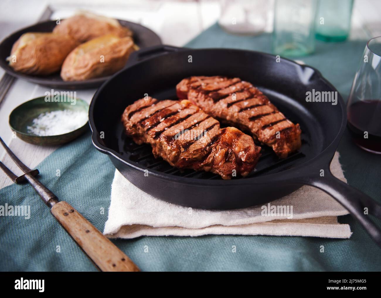 Zwei Grass Fed Steaks in einer gusseisernen Pfanne mit gebackenen Kartoffeln im Hintergrund Stockfoto