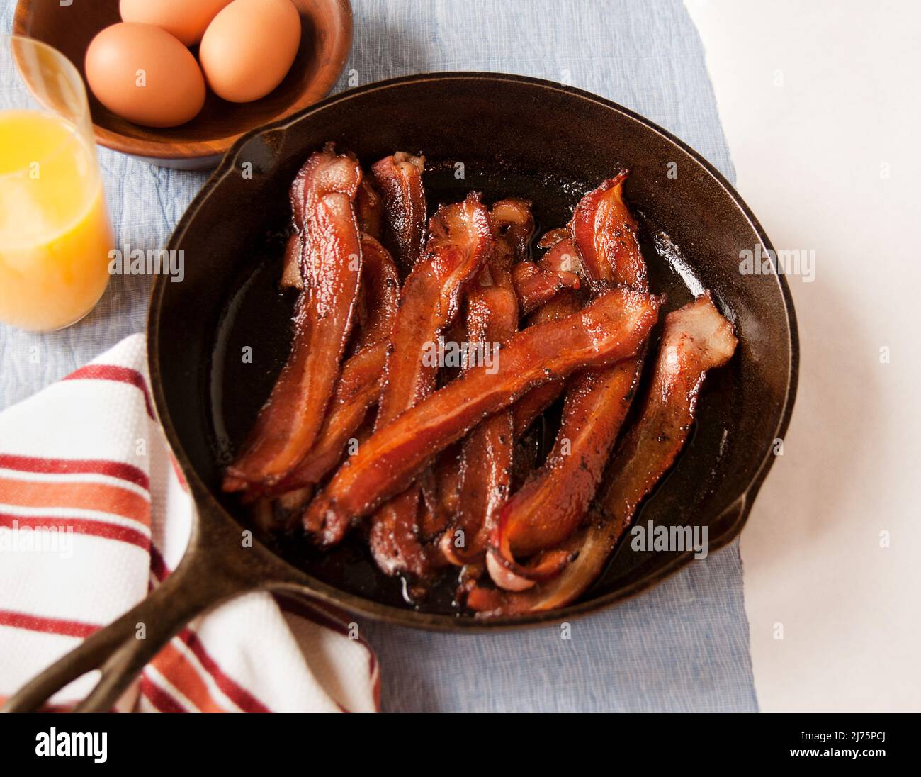 Knusprige Bacon-Streifen in einer gusseisernen Pfanne; ein Glas Orangensaft und eine Schüssel Eier Stockfoto