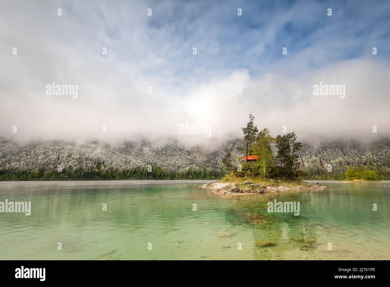 Eine kleine einsame Insel mit Blockhütte und rotem Dach, inmitten des schönen Eibsee unterhalb der Zugspitze in den bayerischen Alpen des Wettersteingebirges während der Eisheiligen - frischer Schnee im Hintergrund auf der Wald- und Wolkenatmosphäre. Stockfoto