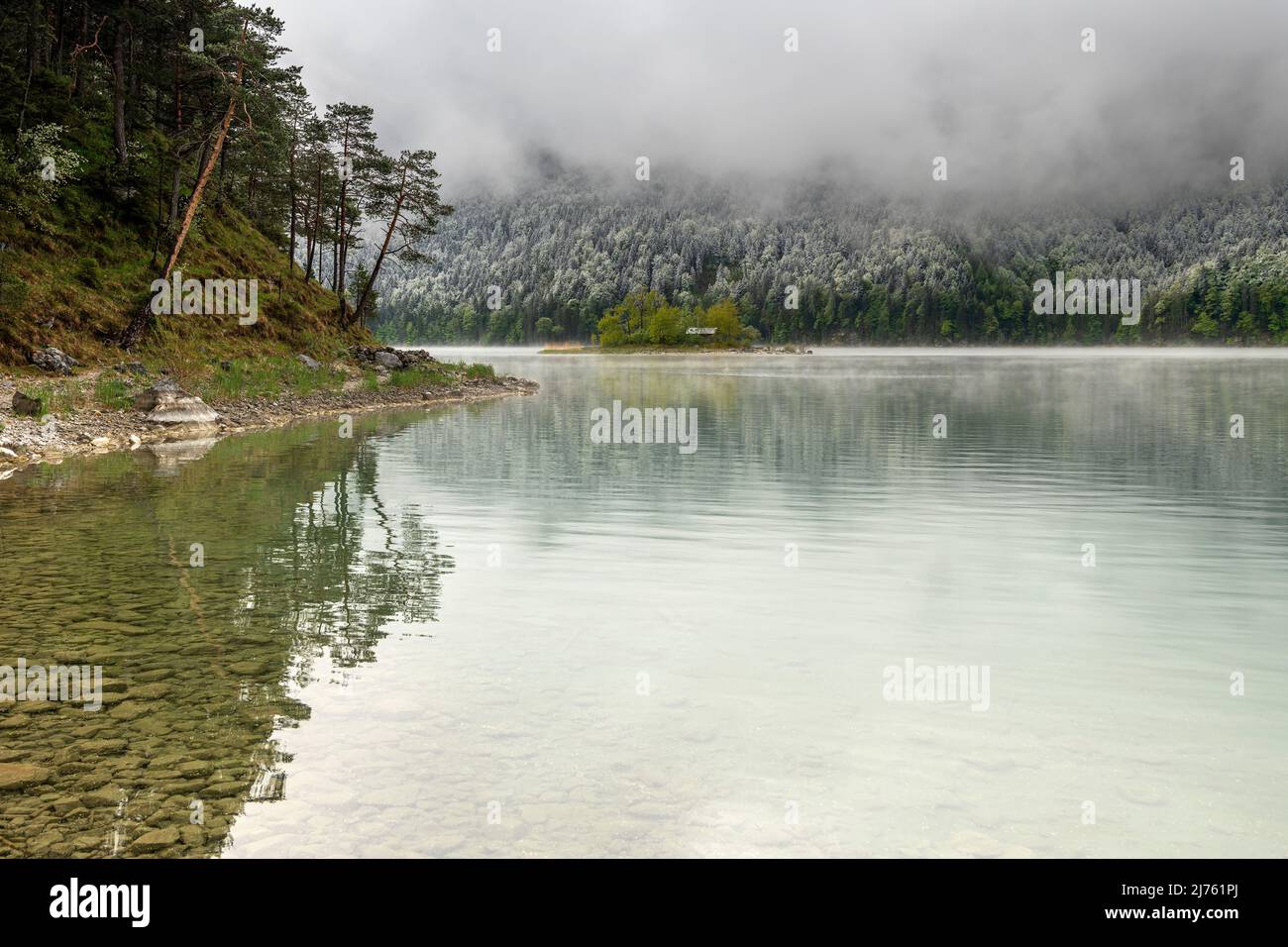 Kleine grüne Insel im Eibsee, unterhalb der Zugspitze in den bayerischen Alpen des Wettersteingebirges während der Eisheiligen im Frühjahr. Im Hintergrund Schnee auf dem frischen Grün des Waldes und dichten Regenwolken. Stockfoto