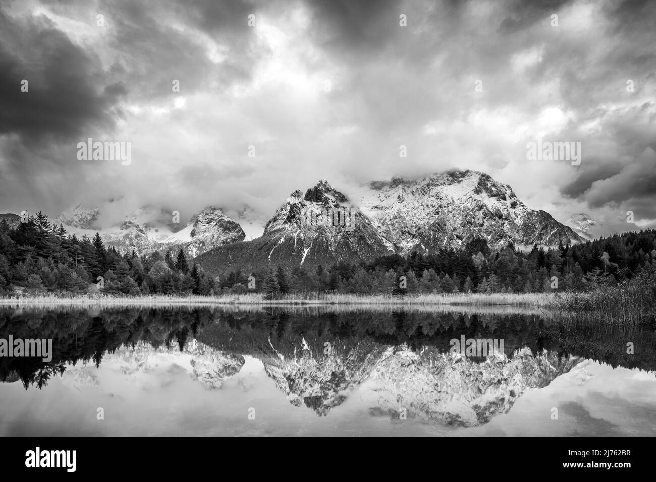 Die Karwendelberge spiegeln sich im Luttensee bei Mittenwald. Frischer Schnee und atmosphärische Wolken umrahmen die Berge. Die schwarz-weiße Umwandlung lässt den Bergrücken massiv wirken. Stockfoto