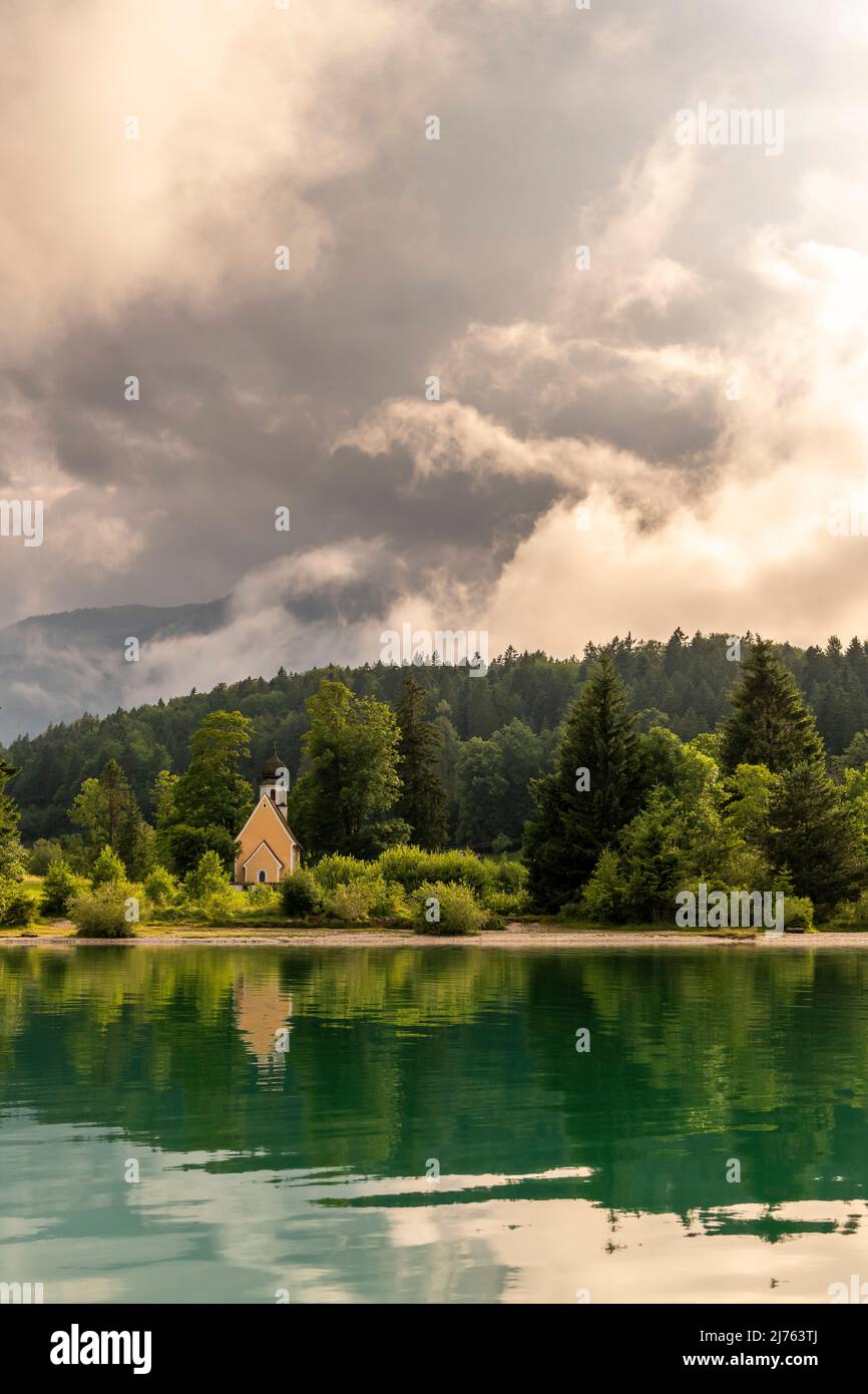 Die Kapelle St. Margareth auf der kleinen Halbinsel Zwergern im Walchensee, fotografiert von einem Boot aus. Im Hintergrund der Simetsberg in dichten Wolken kurz vor Sonnenuntergang, im Vordergrund türkisfarbenes Wasser mit Spiegelung. Stockfoto