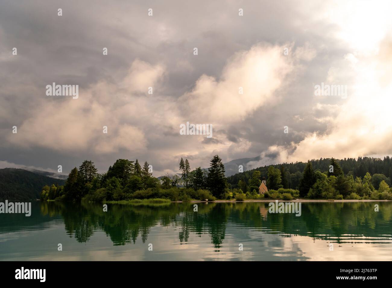 Die Kapelle St. Margareth auf der kleinen Halbinsel Zwergern im Walchensee, fotografiert von einem Boot aus. Im Hintergrund der Simetsberg in dichten Wolken kurz vor Sonnenuntergang, im Vordergrund türkisfarbenes Wasser mit Spiegelung. Stockfoto