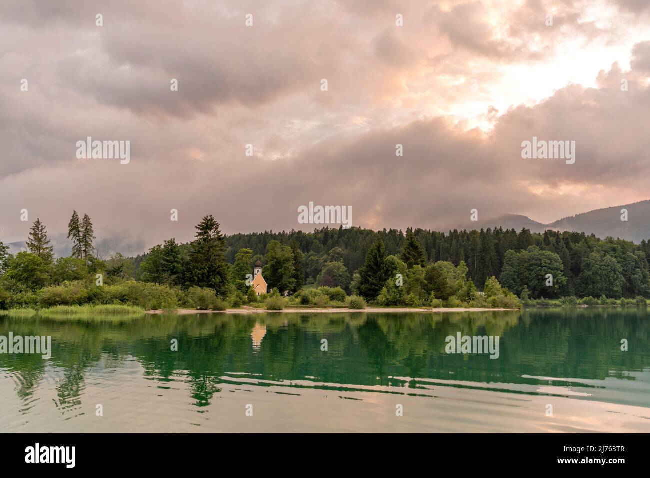 Die Kapelle St. Margareth auf der kleinen Halbinsel Zwergern im Walchensee, fotografiert von einem Boot aus. Im Hintergrund der Simetsberg in dichten Wolken kurz vor Sonnenuntergang, im Vordergrund türkisfarbenes Wasser mit Spiegelung. Stockfoto