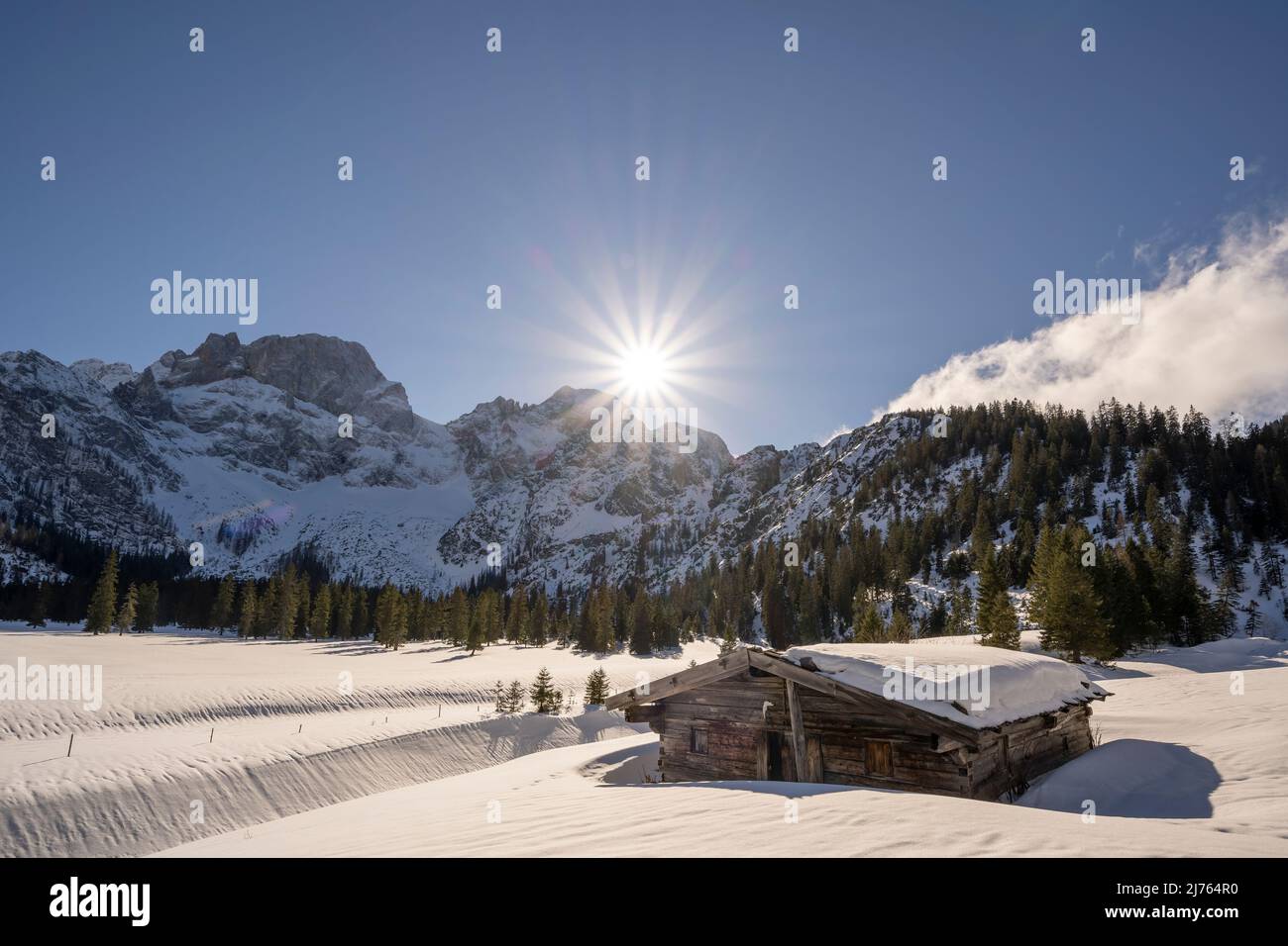 Eine kleine alte Holzhütte im Rhonetal, bei Hinterriss im Karwendel, einem Teil der Alpen in Österreich im Winter mit Schnee. Die Sonne ist tief über den Bergen mit der östlichen Karwendelspitze, mit einem überwiegend blauen Himmel und schönem Wetter, die Hütte wirft ihren Schatten auf den Betrachter, im Hintergrund die Landschaft im Schnee. Stockfoto