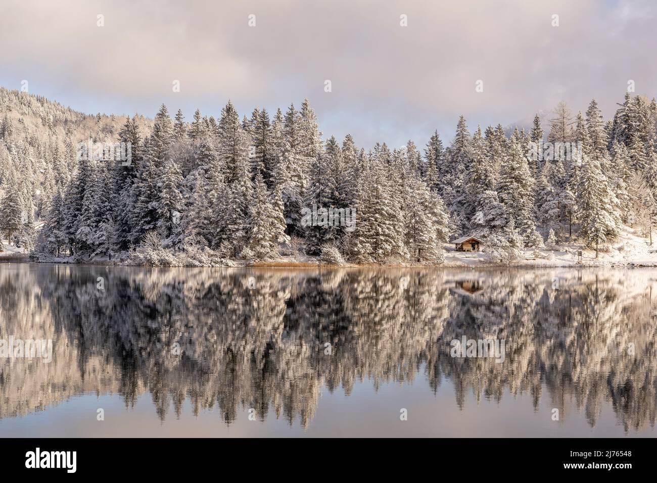Die kleine Hütte gegenüber der Wasserwacht am Lautersee bei Schnee und leichten Wolken an einem sonnigen Wintertag. Das Seeufer spiegelte sich im Wasser des kleinen Bergsees wider Stockfoto