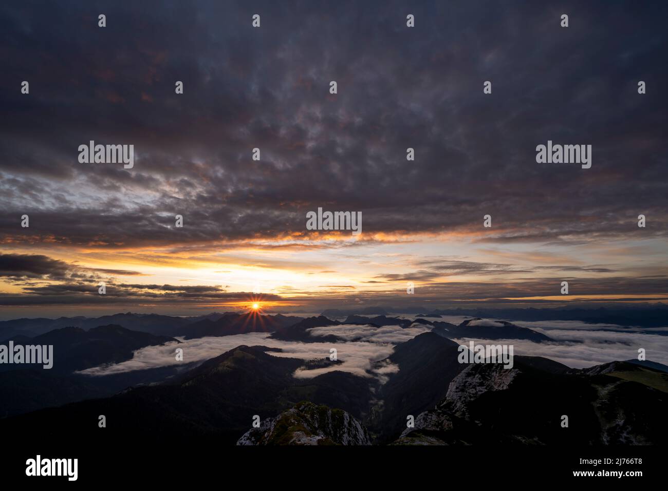 Sonnenaufgang mit Sonnenstern und hellen Wolken, während sich im Tal Nebel und Wolken bewegen. Blick von Guffert (2194m) in Rofan am frühen Morgen. Stockfoto