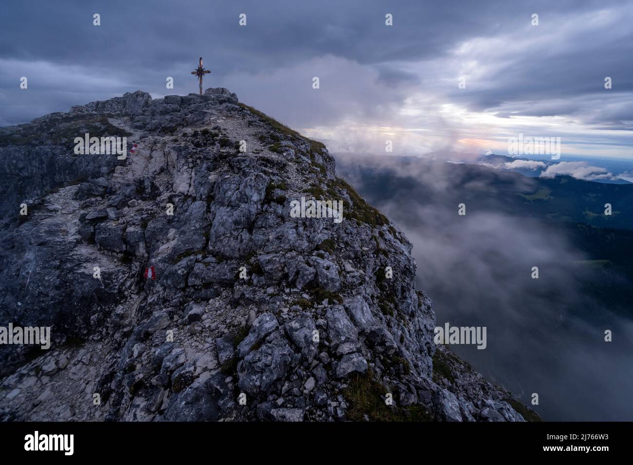 Der Weg zum Gipfel und Gipfelkreuz des Guffert bei schlechtem Wetter und starkem Wind bei Sonnenuntergang. Dichte Wolken und Nebel verdecken einen Großteil der Farben, aber ein leichter farbiger Schein der untergehenden Sonne scheint durch. Stockfoto