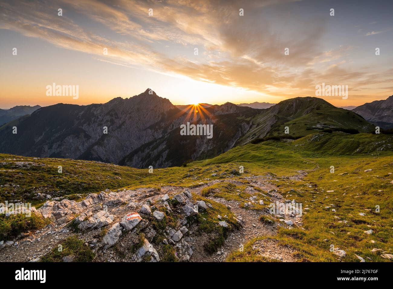 Wanderweg mit Wandermarkierungen, im Hintergrund die Mondscheinspitze mit einem tollen Sonnenstern mit hellen Wolken am sonst blauen Himmel und goldenen, grünen Bergwiesen. Der Weg führt über einen Grat zur Plumsjochhütte von Kompar im Herzen des Karwendels. Stockfoto