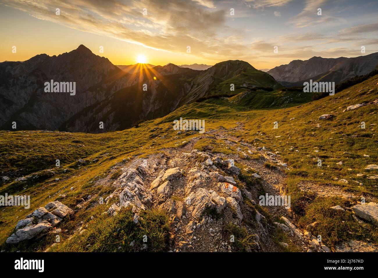 Wanderweg mit Wandermarkierungen, im Hintergrund die Mondscheinspitze mit einem tollen Sonnenstern mit hellen Wolken am sonst blauen Himmel und goldenen, grünen Bergwiesen. Der Weg führt über einen Grat zur Plumsjochhütte von Kompar im Herzen des Karwendels. Stockfoto
