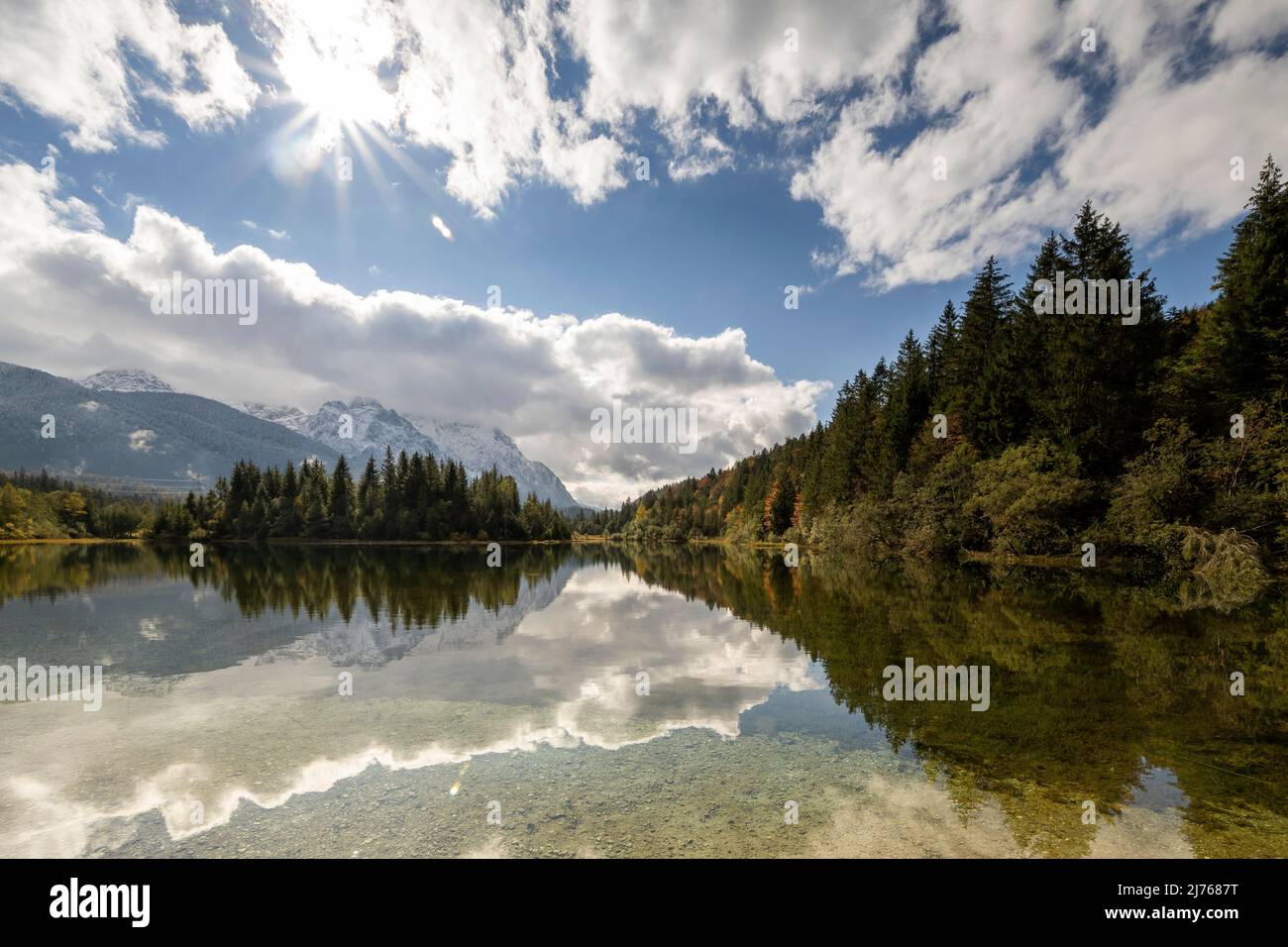 Stausee krün im Herbst mit erstem Schnee am Karwendel. Berge und Wolken spiegeln sich im klaren Wasser des Sees, während sich am Himmel ein Sonnenstern befindet. Stockfoto