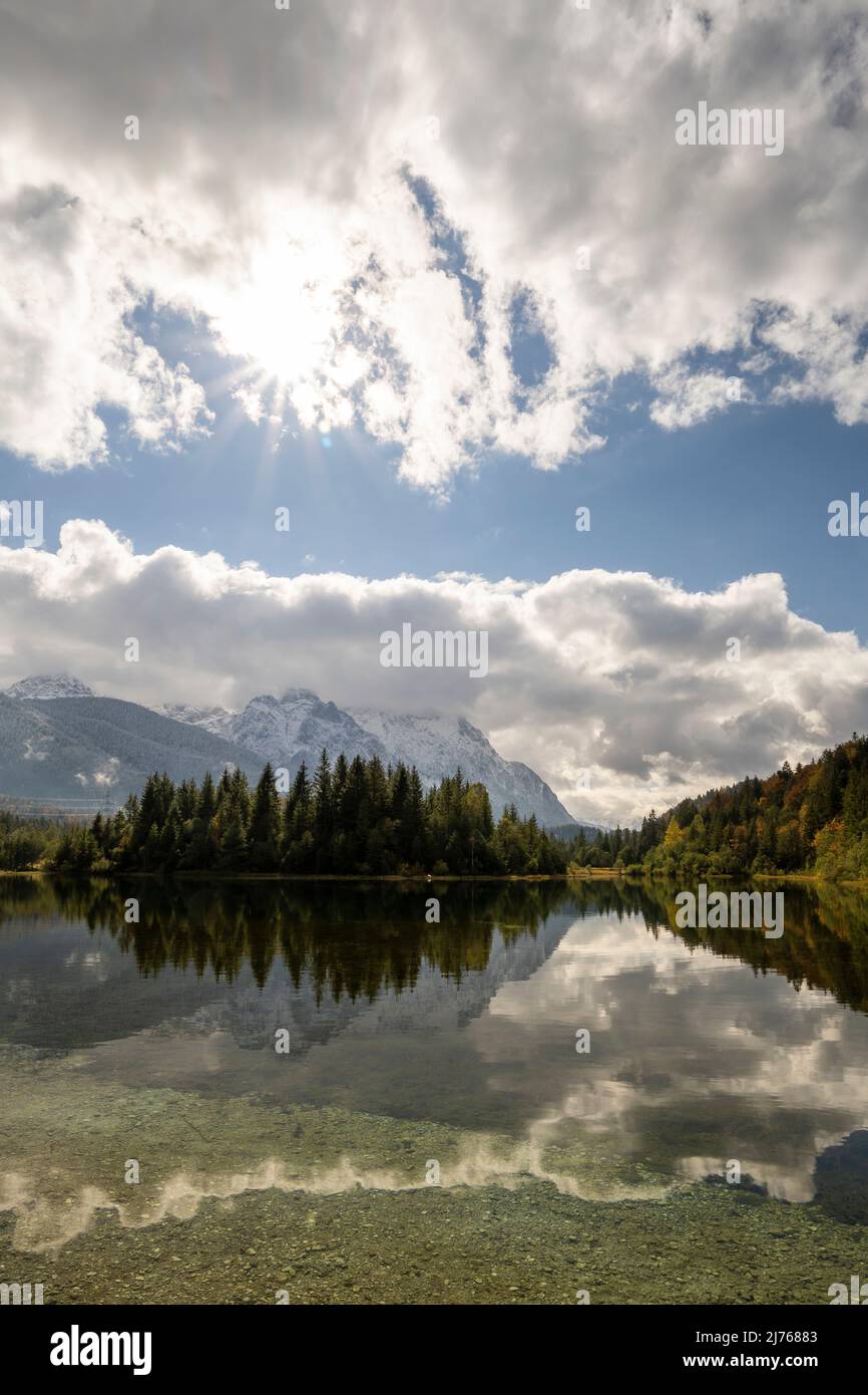 Stausee krün im Herbst mit erstem Schnee am Karwendel. Berge und Wolken spiegeln sich im klaren Wasser des Sees, während sich am Himmel ein Sonnenstern befindet. Stockfoto